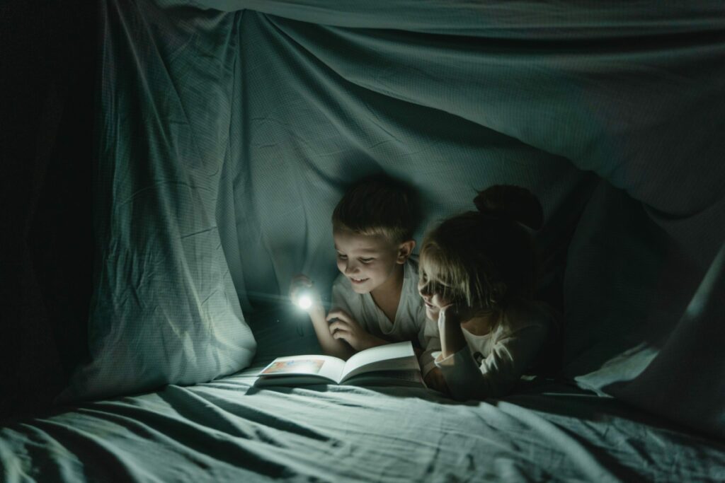Kids reading a book by flashlight under a blanket fort